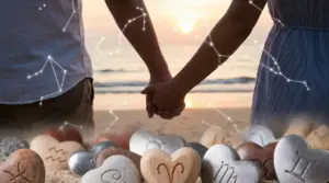 Due persone si tengono per mano sulla spiaggia al tramonto con simboli zodiacali su cuori di pietra